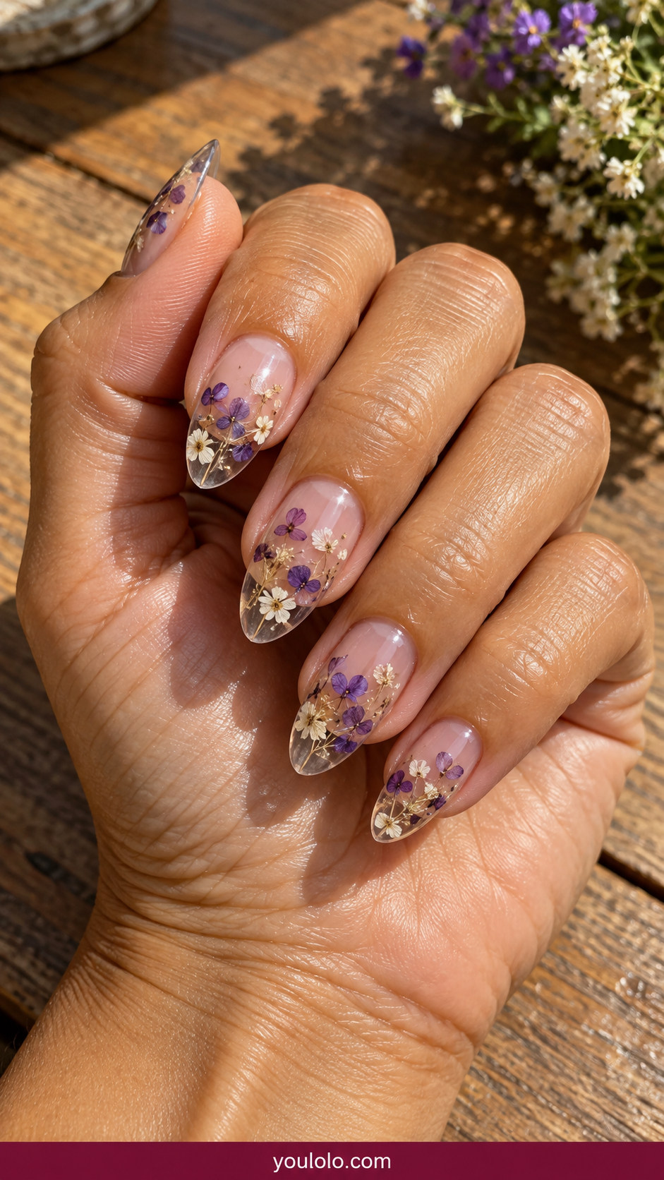 Pressed Flower Nails idea 10: transparent almond tips containing miniature dried purple and white petals on an anatomically realistic feminine hand