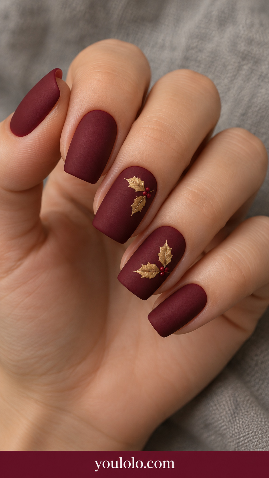 medium soft square matte deep crimson nails with hand-painted fine muted gold holly leaf pair on two accents, tiny burgundy berries, remaining plain matte crimson on a naturally relaxed adult hand