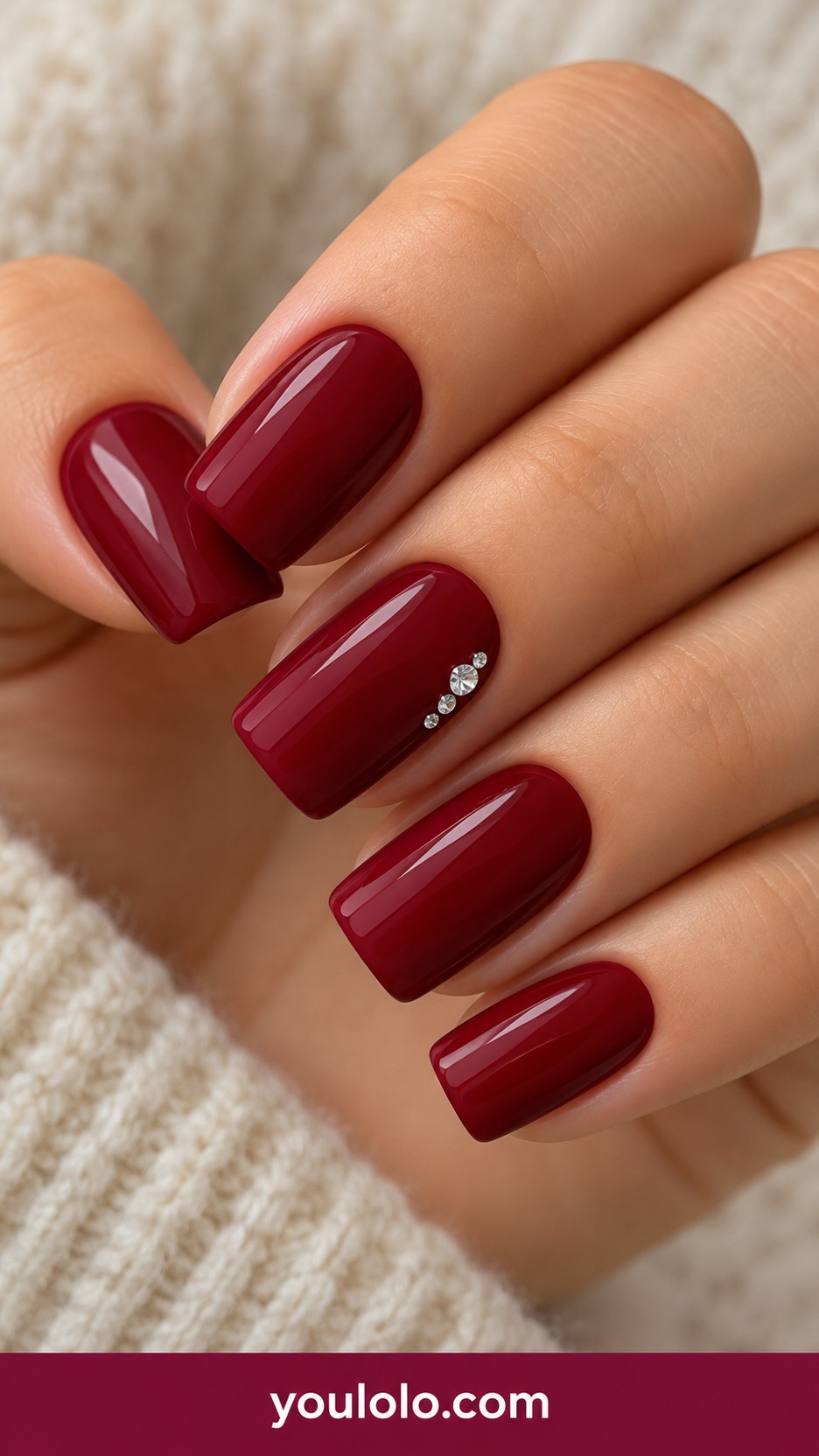 Medium tapered square ruby red nails one accent with three tiny clear crystals at one corner on a naturally curled young adult hand