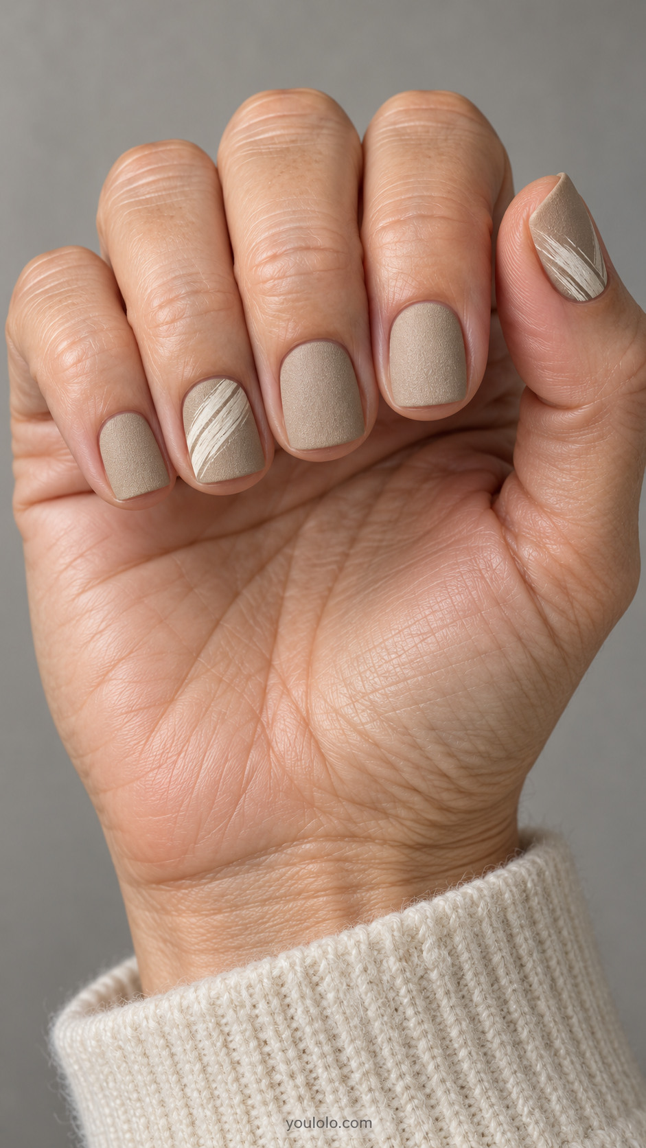 Short square nails: matte sandstone beige nails with irregular narrow ivory dry-brush marks diagonally across ring and thumb, subtle painted texture.