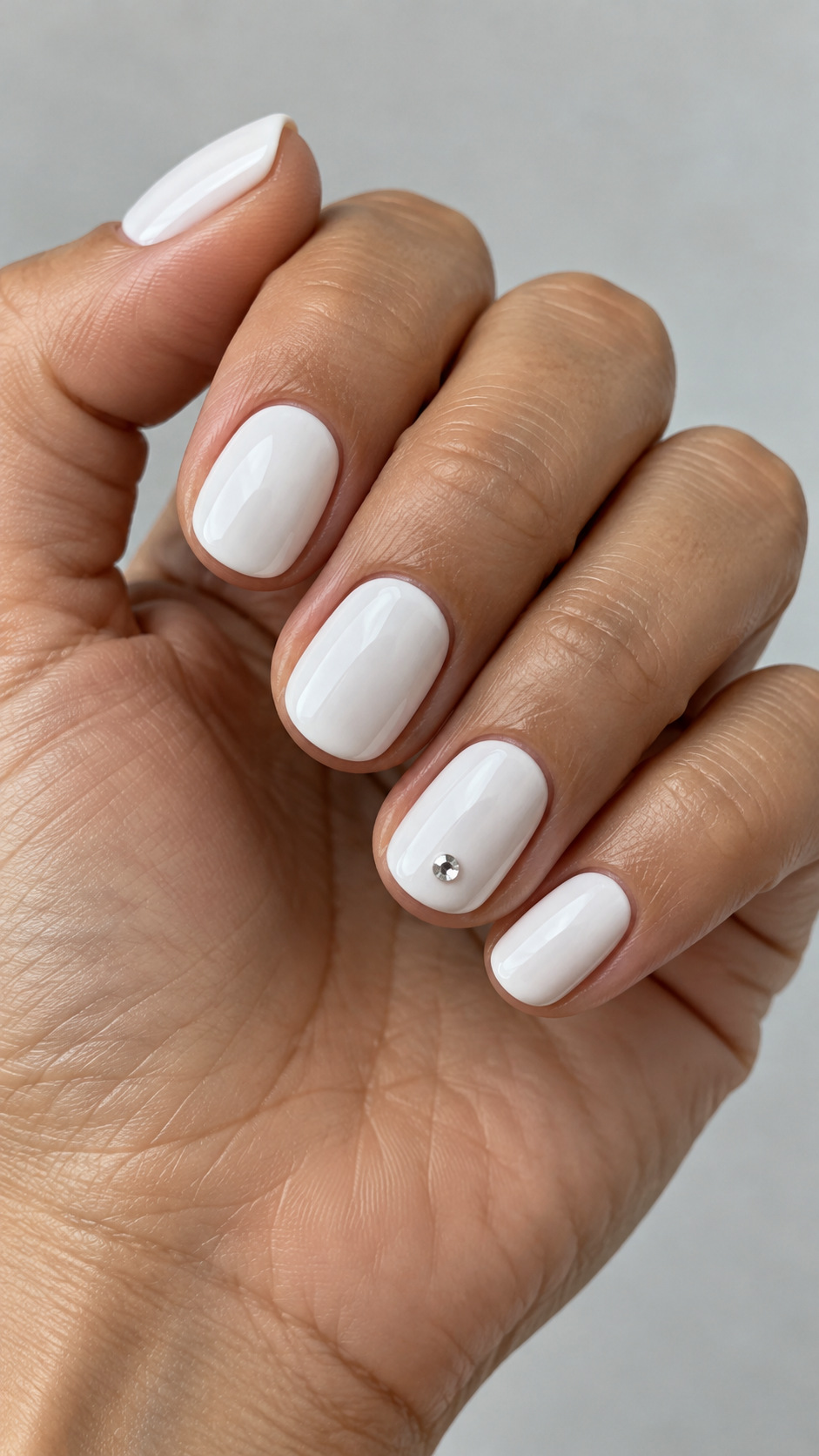 Short White Nails with One Crystal: Short rounded white nails with one small clear crystal on the ring finger on a naturally posed adult hand