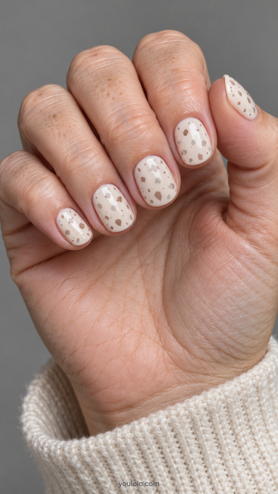 Short round nails: warm ivory nails with sparse tiny irregular cocoa taupe and pale gray terrazzo chips painted flat, no glitter.