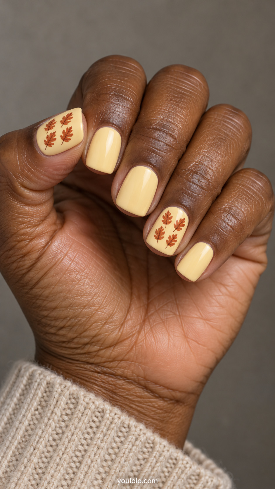 Short square nails with creamy pale yellow nails; ring and thumb have four tiny terracotta oak leaf stamps in a precise two by two tile arrangement; other nails plain.