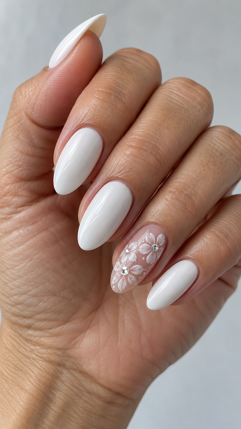 White Flowers with Crystal Centers: White almond nails with painted white flowers and crystal centers on a nude ring nail on a naturally posed adult hand