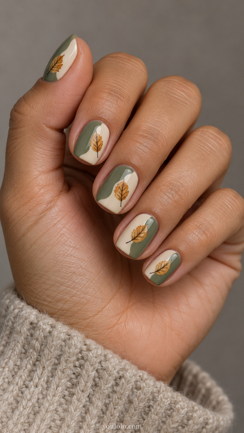 Short squoval nails with each nail divided into two irregular flat color blocks sage green and warm ivory; one tiny ochre beech leaf positioned over join, fine brown veins.