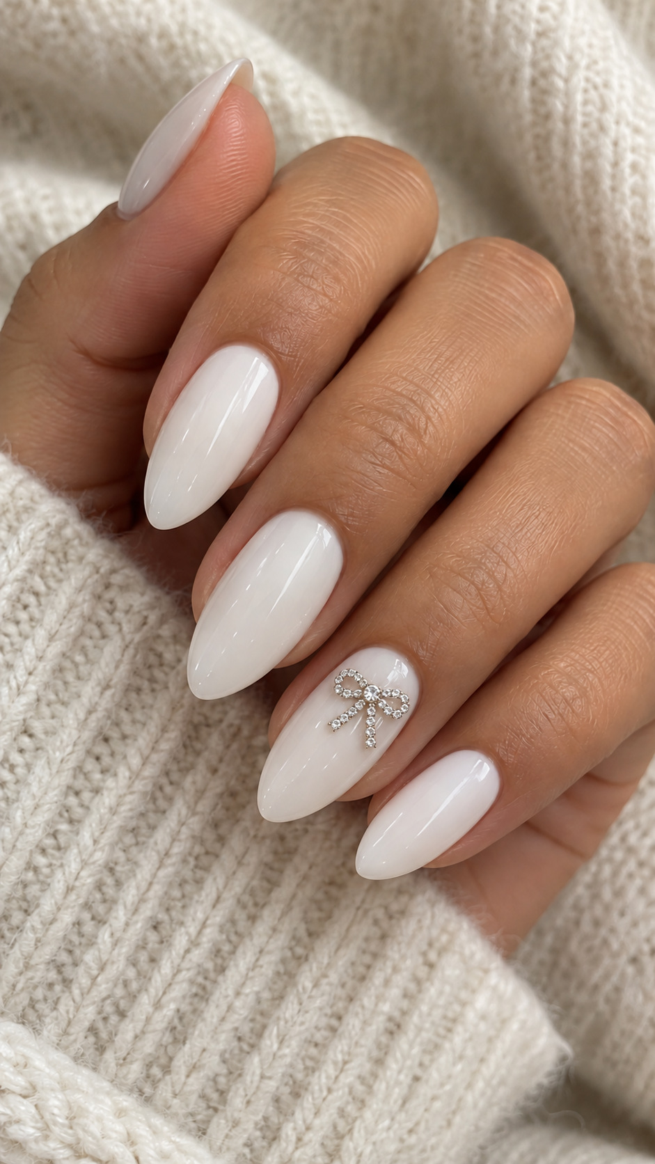 White Nails with a Crystal Bow: White almond nails with a small rhinestone ribbon bow on the ring finger on a naturally posed adult hand