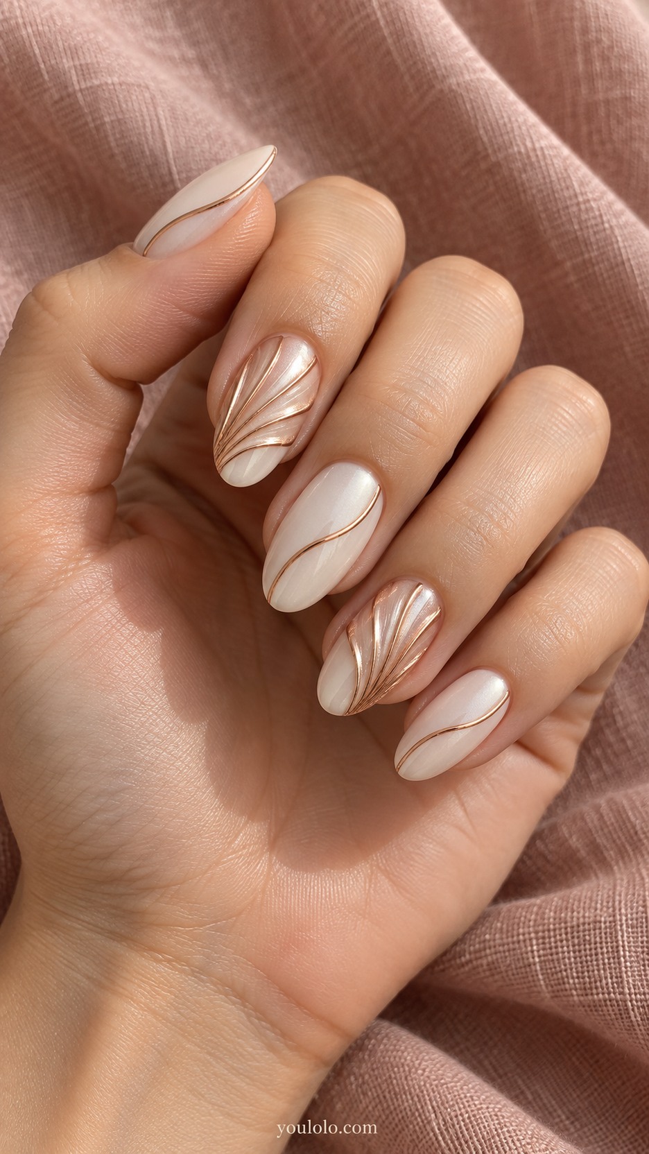 Rose-Gold Folded Fan Nails — Medium almond nails, soft ivory satin base, two accent nails with rose-gold low sculpted fan pleats spreading from one lower side diagonally across nail like folded metallic paper, remaining nails one curved rose-gold line