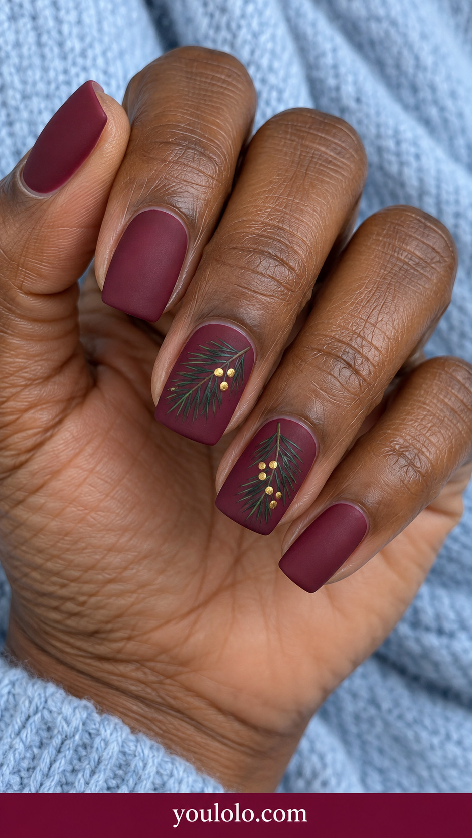 medium soft square matte burgundy nails with delicate flat painted dark forest green FIR NEEDLE twigs and tiny gold berry dots on two accent nails, thin straight short needles rather than broad leaves, remaining plain matte burgundy on a naturally relaxed adult hand