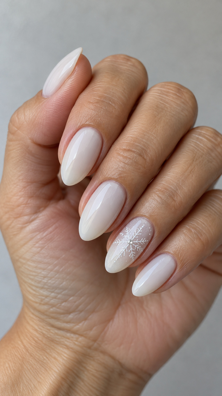 White Snowflakes with Crystal Centers: Milky almond nails with a white snowflake and tiny crystal center on one finger on a naturally posed adult hand
