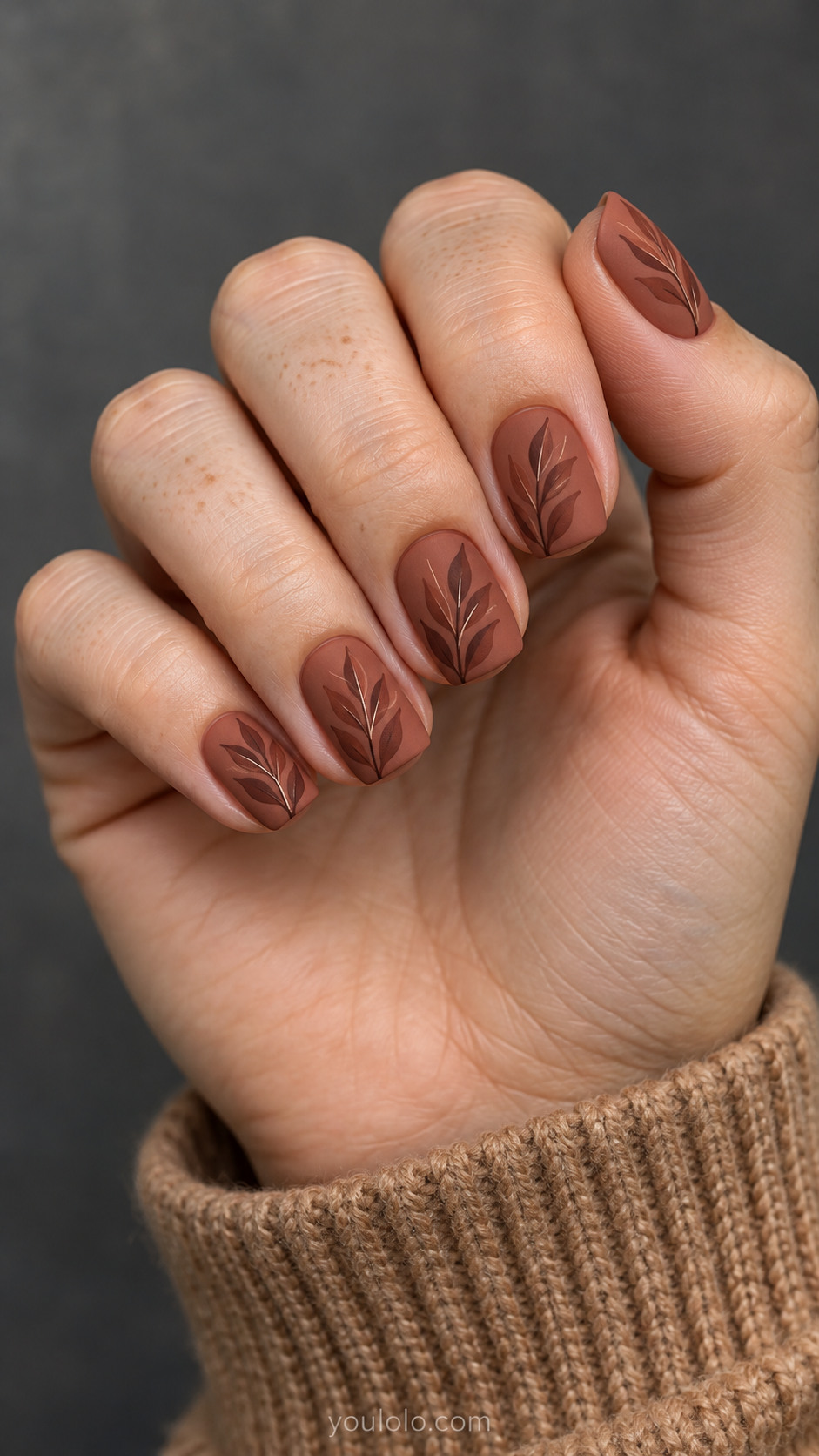 Short matte terracotta squoval nails with darker painted leaf sprigs and fine lighter stems.