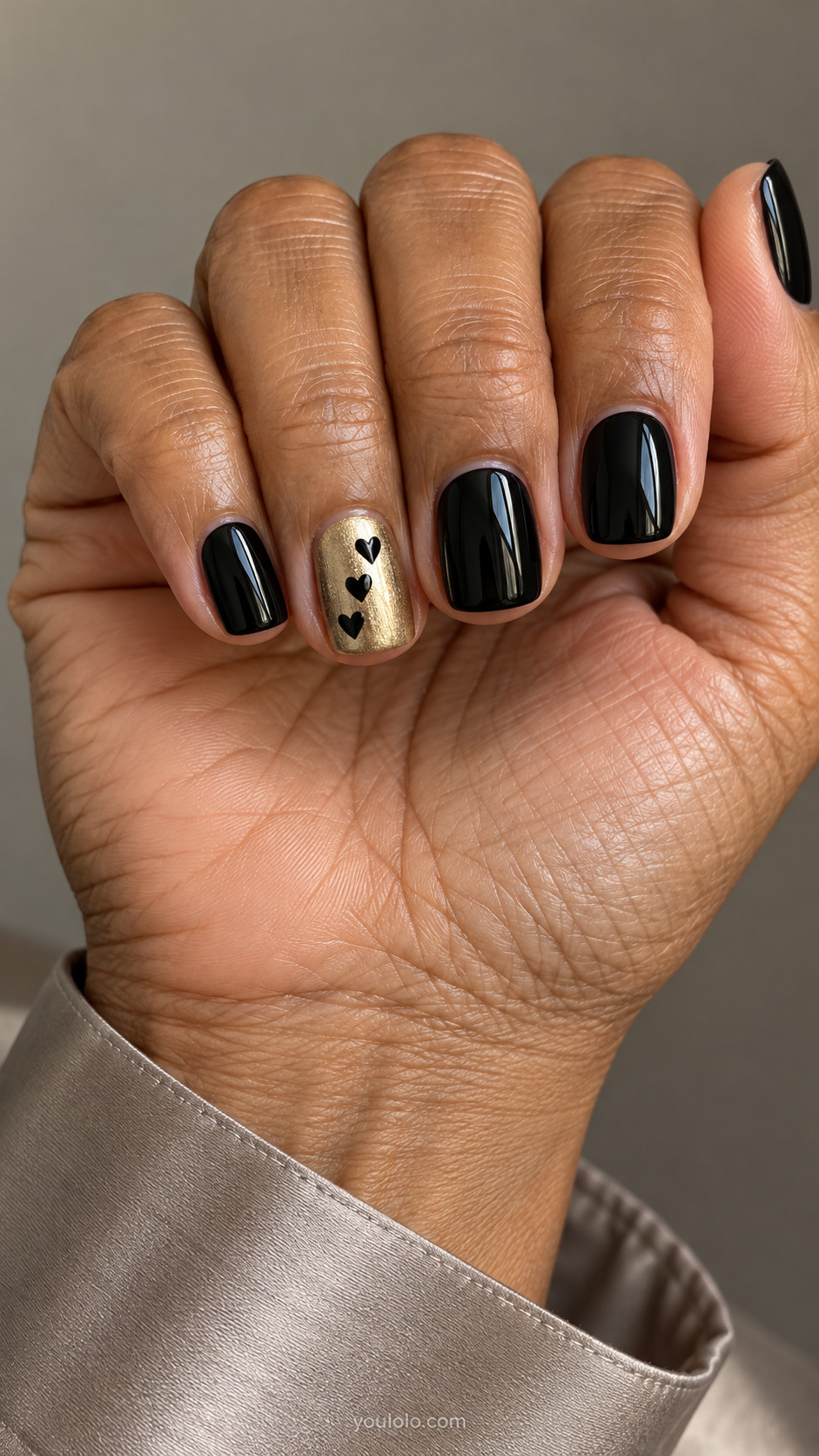 Short black nails with one metallic gold accent decorated with three black hearts