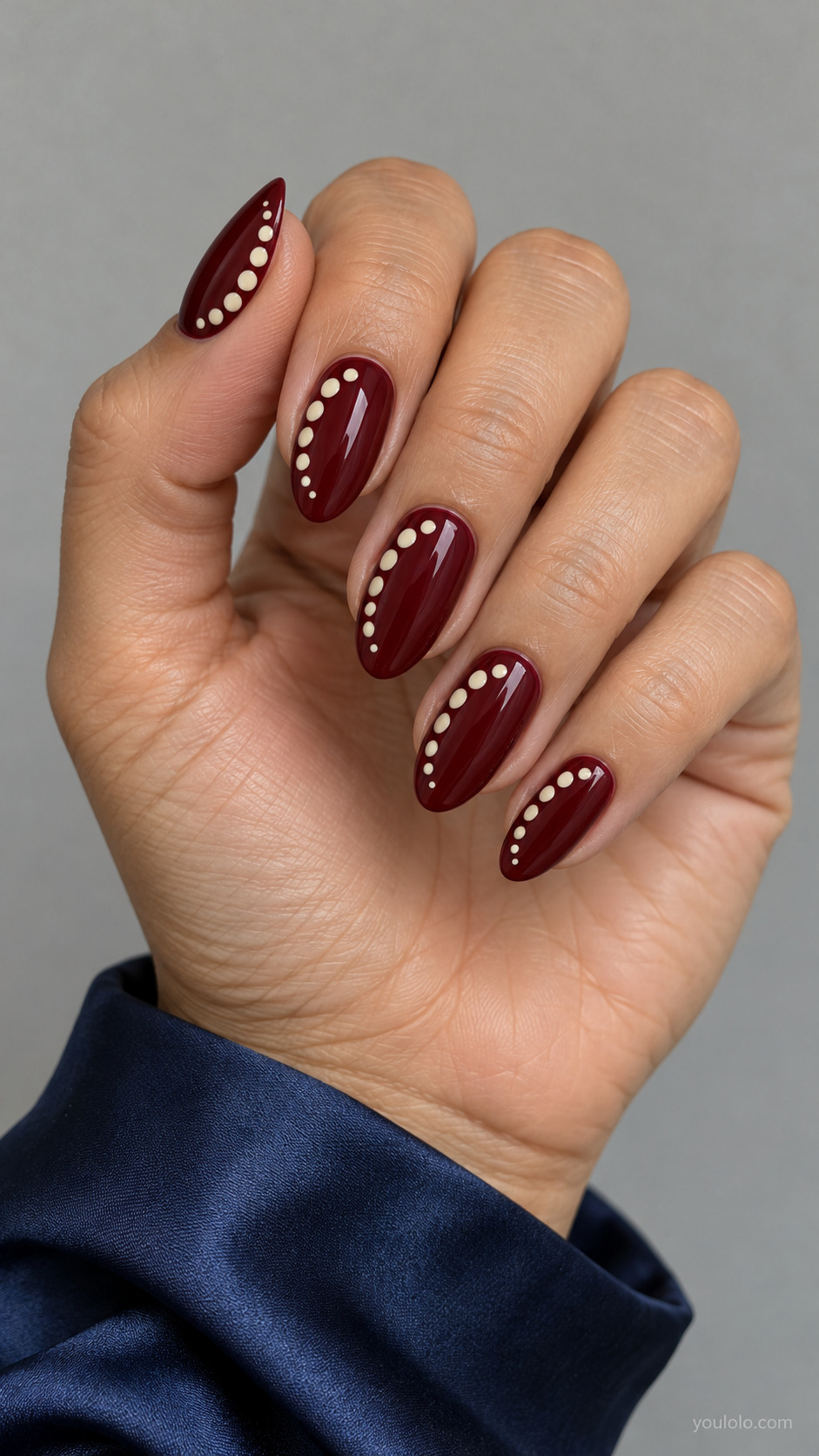 Medium almond nails full burgundy, ivory dots concentrated along one side of each nail in curved crescent arrangement, other side empty.
