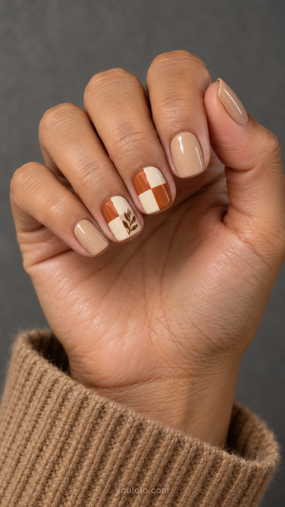 Short beige square nails with two orange-and-ivory checker accents and a brown leaf sprig on one.