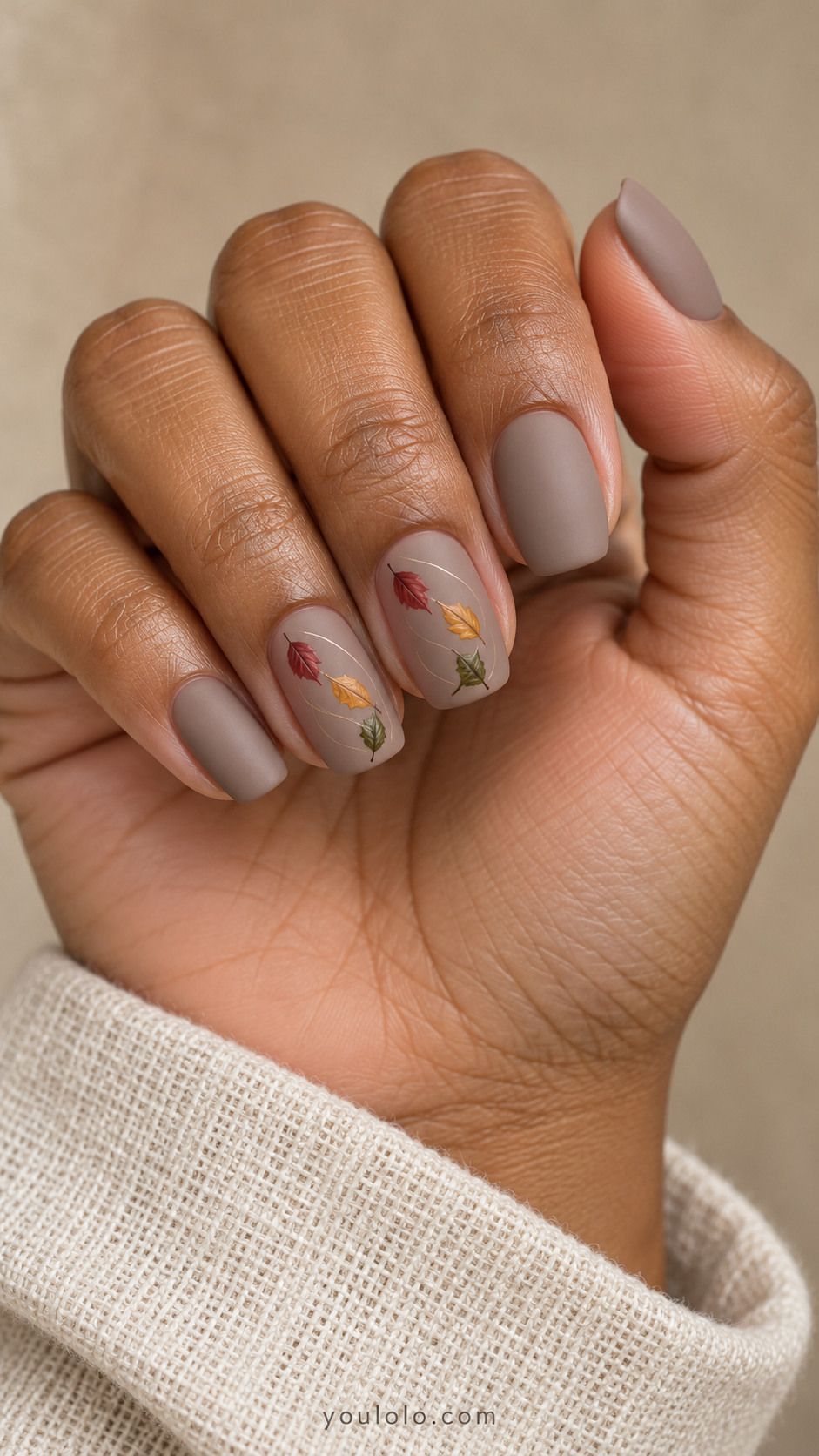 Medium rounded square nails with matte taupe nails; three tiny handpainted leaves red amber and olive tumble diagonally across each middle and ring nail with very fine curved wind line; others plain taupe.